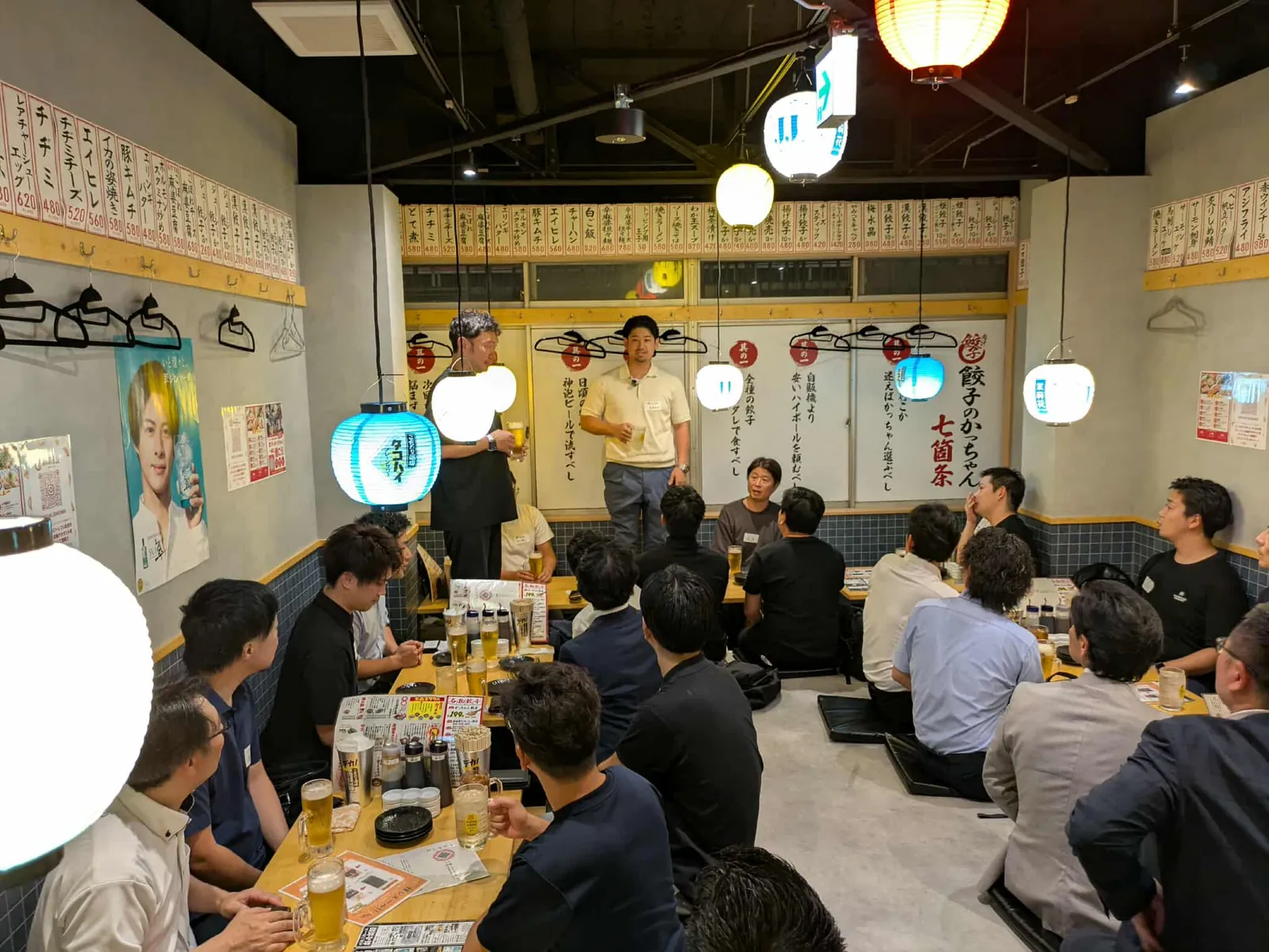 提灯の下がる居酒屋で、前に立って話す2人と、座敷の席で聞く参加者