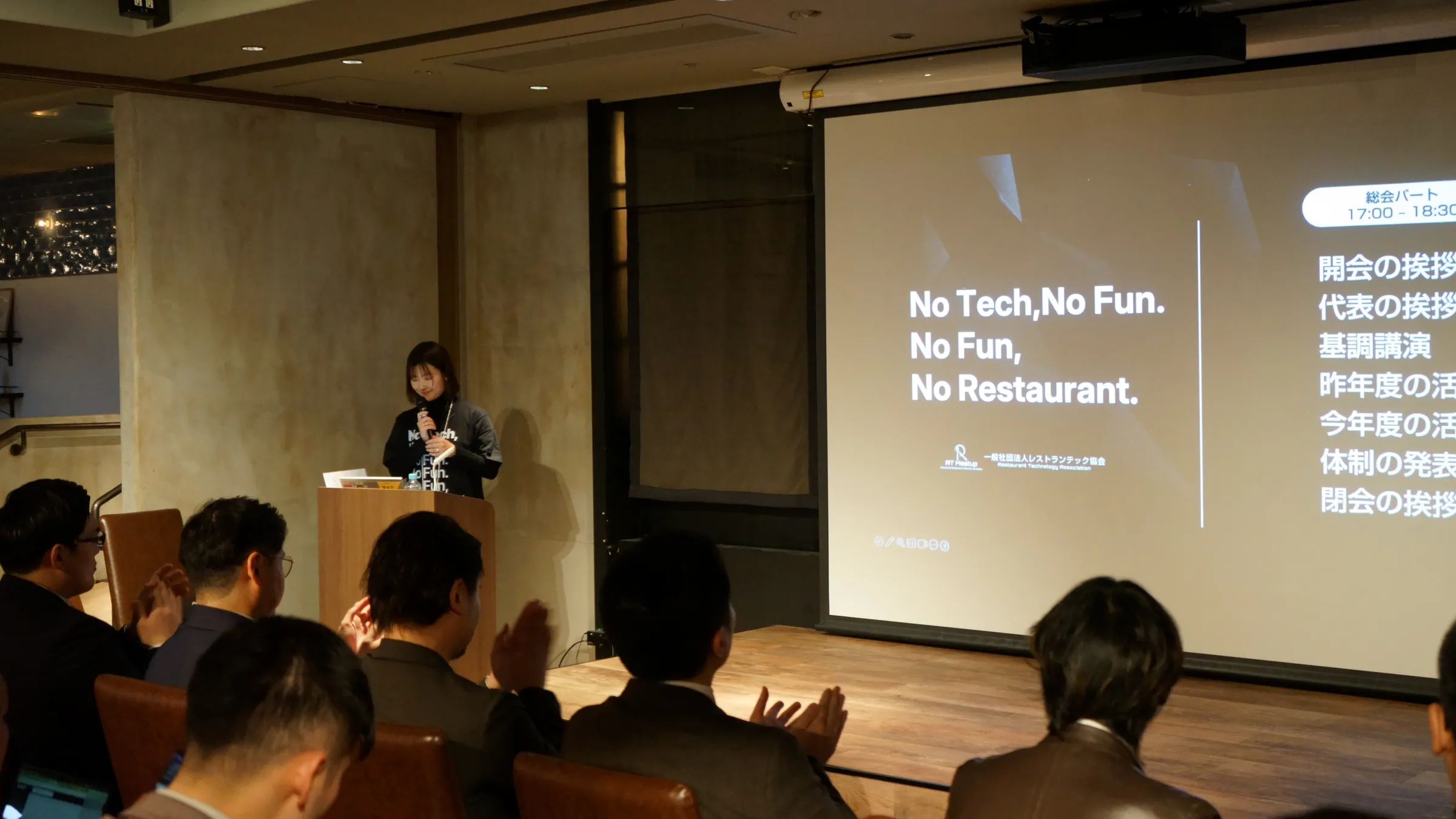 2025年3月の総会・懇親会で、「No Tech, No Fun, No Restaurant.」のスライドの前で、話を聞く参加者